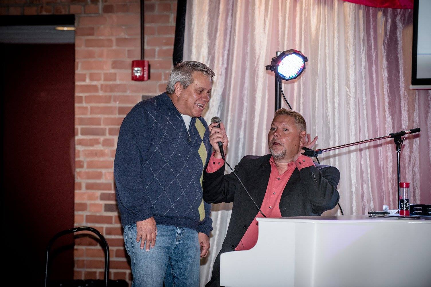 A man playing piano with another man holding a microphone, brick wall background.
