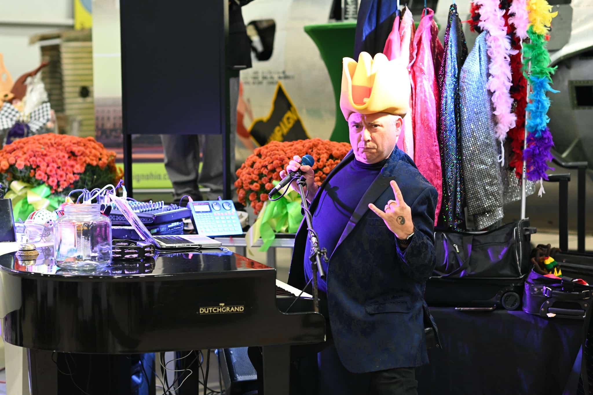 A person with a yellow hat sings into a microphone by a piano, making a rock hand gesture.
