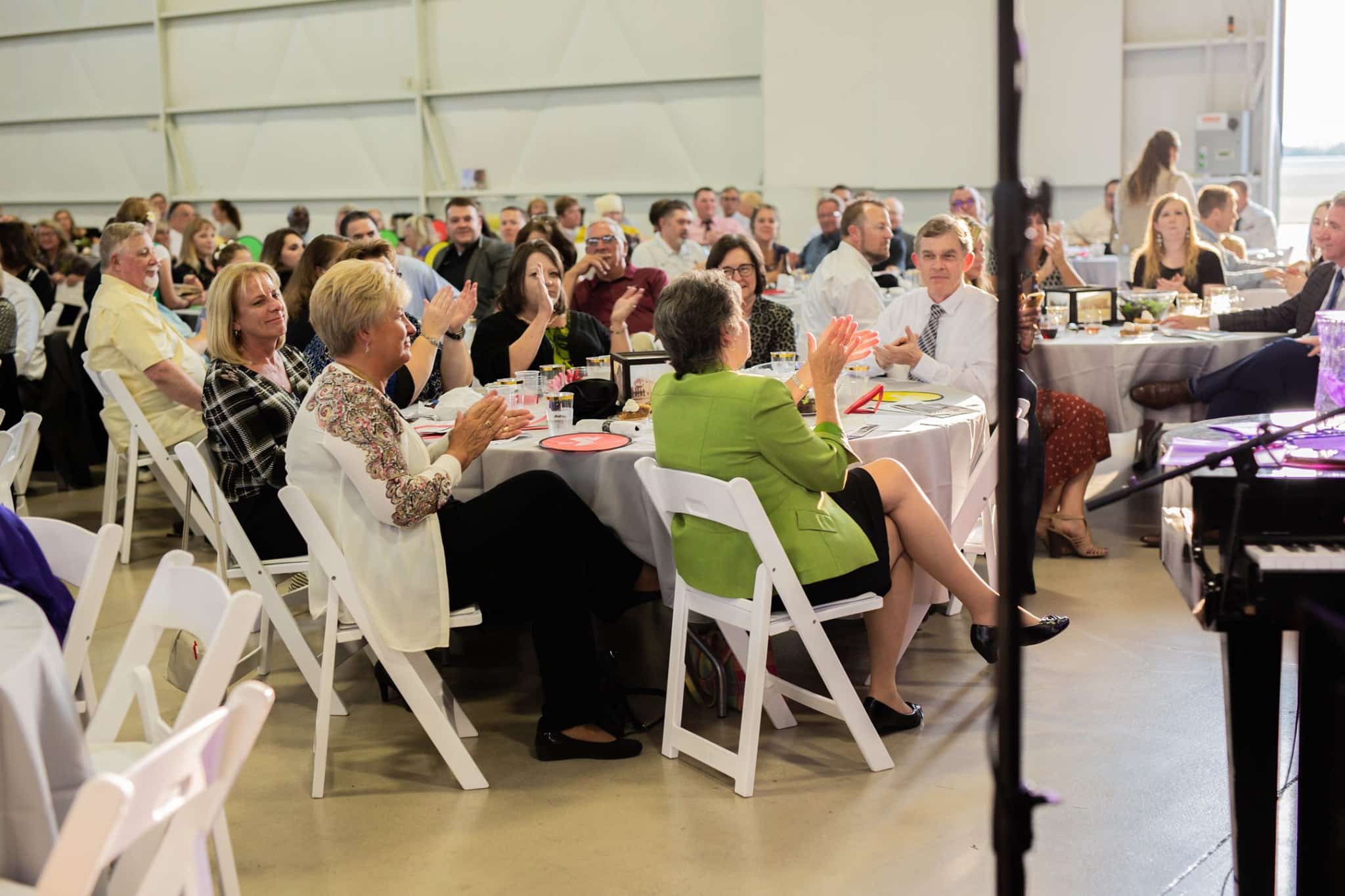 People applauding at a banquet event with round tables and white chairs.