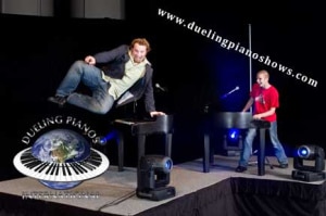 Two men performing a high-energy dueling piano show on stage.