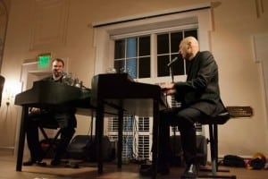 Two musicians perform at pianos in an elegant room with large windows.