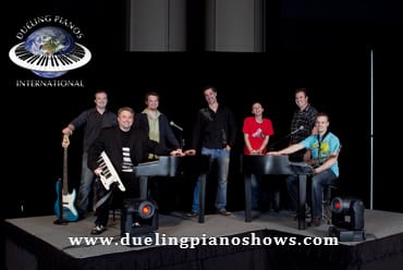 Band members performing at Dueling Pianos International event with instruments on stage.