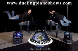 Two people mid-air over pianos on a stage with duelingpianoshows.com banner.