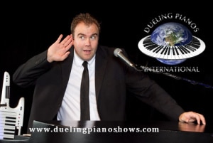 Man at piano with microphone, hand to ear, promoting Dueling Pianos International.