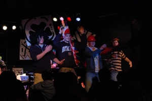 Four men on stage in casual clothes and hats performing energetically.
