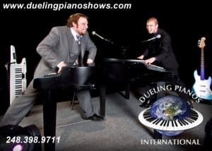 Two men playing pianos on stage for a dueling piano show, with musical instruments nearby.