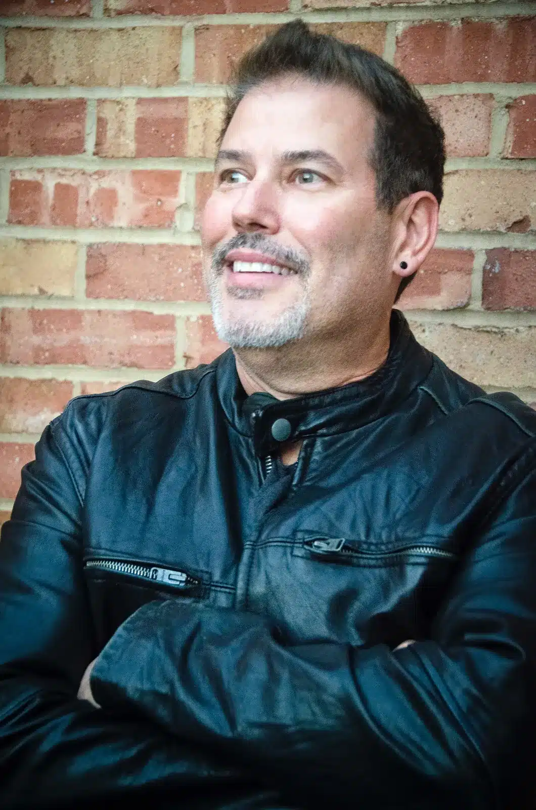 A man in a black leather jacket smiling against a brick wall background.